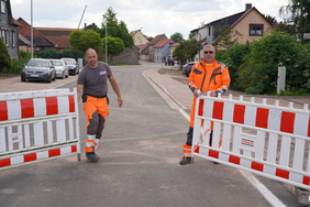 Zu sehen sind zwei Männer, Ringo Frommann und Rainer Kreidel, in Arbeitskleidung. Sie stehen auf einer Straße und öffnen Bauzäune.