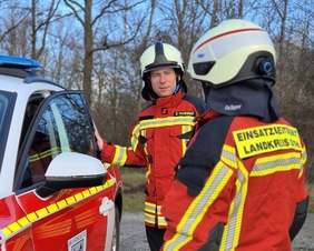 Zu sehen sind zwei Männer in Feuerwehr-Einsatzbekleidung. Sie stehen neben einem Einsatzwagen und sprechen miteinander. 