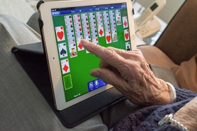 Zu sehen ist die Hand einer älteren Person auf einem Tablet. Auf dem Tablet wird ein Kartenspiel angezeigt.